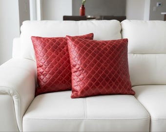 May include: Two red, square throw pillows with a quilted diamond pattern rest on a white leather sofa. The pillows add a pop of color to the neutral-toned living room. The sofa has a modern design.