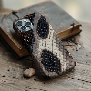 May include: A smartphone case featuring a textured snakeskin pattern in shades of brown, black, and cream. The case has a cutout for a triple-lens camera system on the upper left corner, revealing the metallic blue rings of the camera lenses.