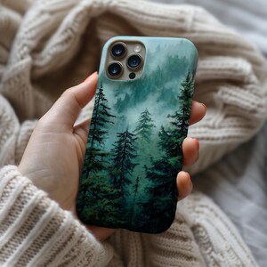 May include: A smartphone case featuring a printed design of a dense, misty forest with tall evergreen trees in shades of deep green and teal. The case has a cutout for a triple-lens camera system on the upper left corner and a smooth finish.