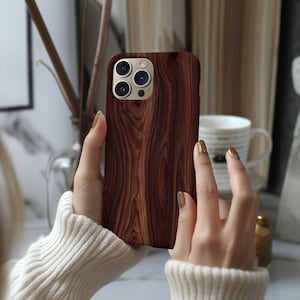 May include: A smartphone case featuring a dark brown wood grain pattern with a smooth finish. The case has a rectangular cutout at the top left corner for a triple camera system and a flash, showing the metallic edges of the phone camera lenses.