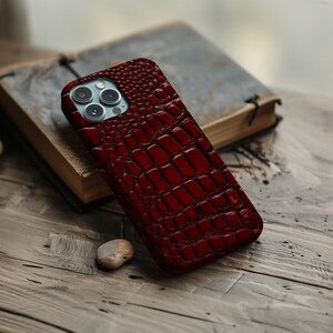 Op de afbeelding: Een dieprode smartphonehoes met een gestructureerd krokodillenleerpatroon. De hoes heeft een uitsparing voor een camera met drie lenzen in de linkerbovenhoek, waarbij de ronde lenzen en een flitsmodule in het frame zichtbaar zijn.