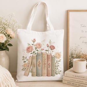Op de afbeelding: Een witte canvas draagtas met een bedrukte illustratie van een rij van vijf boeken in vintage-stijl, versierd met zachtroze en crèmekleurige rozen, groene bladeren en delicate bloemtakjes die boven de ruggen van de boeken zijn geplaatst.