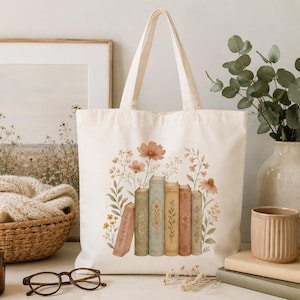 Op de afbeelding: Een crèmekleurige canvas draagtas met een bedrukte illustratie van vijf rechtopstaande boeken in vintage-stijl, omringd door delicate, aquarelachtige wilde bloemen in roze, gele en groene tinten op de voorzijde.