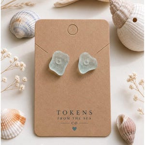 Może przedstawiać: Kolczyki sztyfty z morskiego szkła w kolorze jasnoniebieskim na brązowej karcie z napisem "TOKENS FROM THE SEA CO". Kolczyki mają mały, przezroczysty środek. Muszle i suszone kwiaty otaczają kartę.
