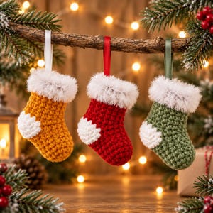 May include: Three small crocheted stockings in mustard yellow, red, and sage green. Each stocking features a white fuzzy cuff, a white heel patch, and a matching satin ribbon loop for hanging. The stockings are suspended from a wooden branch.