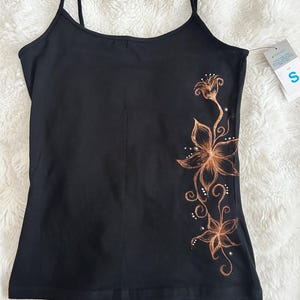 May include: Black camisole top with thin straps. A floral design in brown and white accents decorates the side. A tag with the letter "S" is attached to the top.