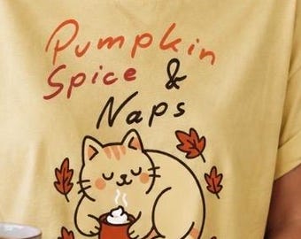 Op de afbeelding: Een lichtgeel T-shirt met korte mouwen met de tekst Pumpkin Spice & Naps in bruine en oranje letters boven een afbeelding van een cartoonkat die een mok vasthoudt, omringd door kleine bruine en oranje herfstbladeren.