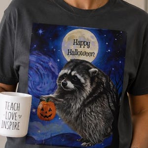 Op de afbeelding: Een antracietgrijs T-shirt met ronde hals en een afbeelding van een wasbeer die een pompoen vasthoudt onder een volle maan met de tekst Happy Halloween. Het shirt wordt gecombineerd met een witte mok met de tekst TEACH LOVE INSPIRE.