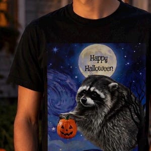 Op de afbeelding: Een zwart t-shirt met korte mouwen en een grafische print van een wasbeer die een kleine oranje pompoen vasthoudt. De achtergrond toont een nachtelijke hemel met een grote volle maan en de tekst Happy Halloween in zwarte letters.