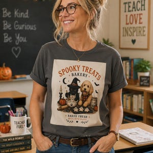 May include: A charcoal gray t-shirt featuring a graphic print of a black cat wearing a witch hat and a golden doodle dog baking. The text reads Spooky Treats Bakery, Baked Fresh, and Made With Magic, accompanied by illustrations of ghosts and pumpkins.