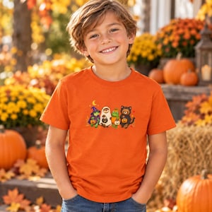 May include: An orange short-sleeved t-shirt featuring a graphic print of four small animals dressed in Halloween costumes, including a frog as a witch, a sloth as a ghost, a frog as a pumpkin, and a bear as a black cat, all holding small pumpkins.