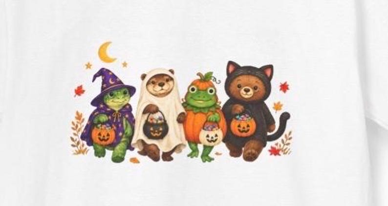 May include: A white t-shirt featuring a colourful illustration of four animals dressed in Halloween costumes: a turtle in a purple wizard hat, an otter in a ghost sheet, a frog in a pumpkin suit, and a bear in a black cat costume, all holding jack-o'-lantern candy buckets.