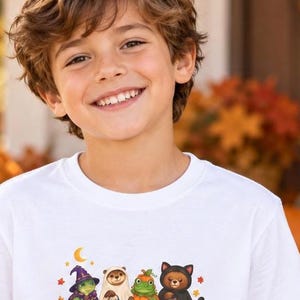 May include: A white t-shirt featuring a graphic print of four small animals dressed in Halloween costumes. The illustration includes a turtle in a witch hat, an otter in a ghost sheet, a frog in a pumpkin suit, and a bear in a black cat costume.