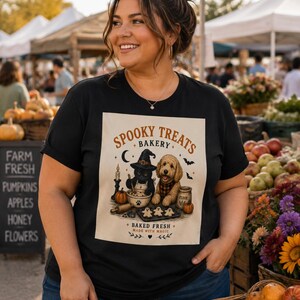 May include: A black short-sleeve t-shirt featuring a graphic print with the text "SPOOKY TREATS BAKERY" and "BAKED FRESH MADE WITH MAGIC". The illustration shows a black cat in a witch hat and a golden dog baking cookies with pumpkins.