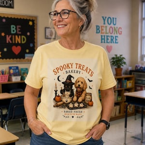 May include: A pale yellow t-shirt featuring a graphic print of a black cat in a witch hat and a golden doodle dog baking cookies. The text reads Spooky Treats Bakery, Baked Fresh, and Made With Magic, accompanied by pumpkins and bats.
