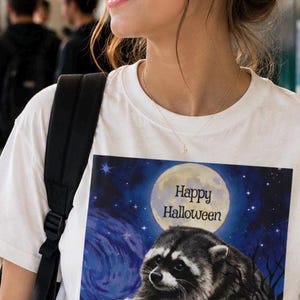 Op de afbeelding: Een wit t-shirt met een grafische print van een wasbeer die een kleine oranje pompoen vasthoudt. De achtergrond toont een sterrenhemel met een grote volle maan en de tekst Happy Halloween in zwarte letters boven de wasbeer.