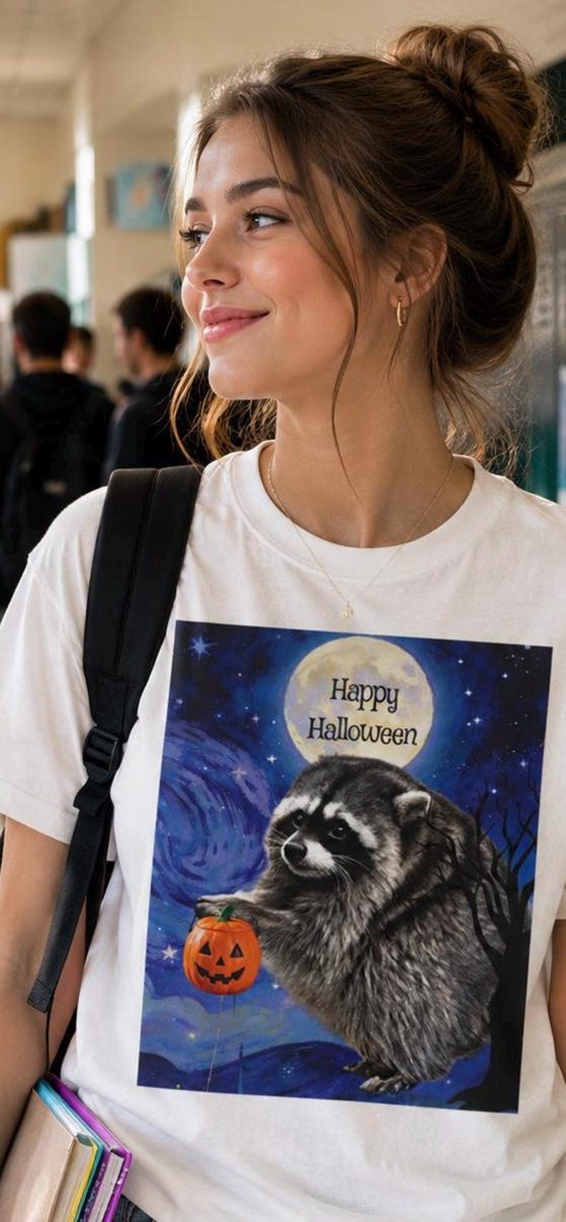 Op de afbeelding: Een wit t-shirt met een grafische print van een wasbeer die een kleine oranje pompoen vasthoudt. De achtergrond toont een sterrenhemel met een grote volle maan en de tekst Happy Halloween in zwarte letters boven de wasbeer.