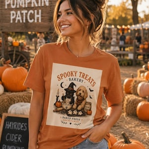 May include: A burnt orange t-shirt featuring a graphic print of a black cat wearing a witch hat and a golden dog baking cookies. The text reads Spooky Treats Bakery, Baked Fresh, and Made With Magic, accompanied by pumpkins and bats.