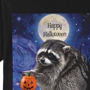 Op de afbeelding: Een zwart t-shirt met een afbeelding van een wasbeer die een kleine oranje pompoen vasthoudt tegen een donkerblauwe nachtelijke hemel met een volle maan en sterren. De tekst "Happy Halloween" staat in zwarte letters onder de maan gedrukt.