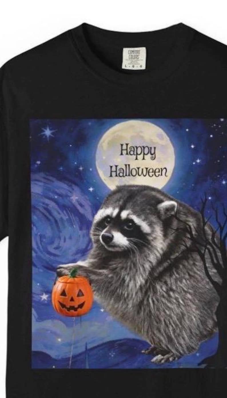 Op de afbeelding: Een zwart t-shirt met een afbeelding van een wasbeer die een kleine oranje pompoen vasthoudt tegen een donkerblauwe nachtelijke hemel met een volle maan en sterren. De tekst "Happy Halloween" staat in zwarte letters onder de maan gedrukt.