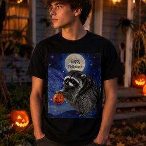 Op de afbeelding: Een zwart t-shirt met een grafische print van een wasbeer die een kleine oranje pompoen vasthoudt. De achtergrond toont een volle maan in een donkerblauwe nachtelijke hemel met de tekst Happy Halloween in zwarte letters onder de maan.
