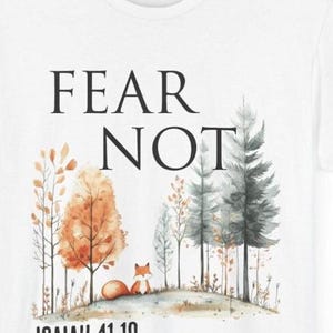 May include: A white t-shirt featuring a watercolor-style graphic of a small orange fox sitting in a forest with orange and grey trees. The text "FEAR NOT" is printed in black above the forest, and "ISAIAH 41:10" is printed in black below the fox.