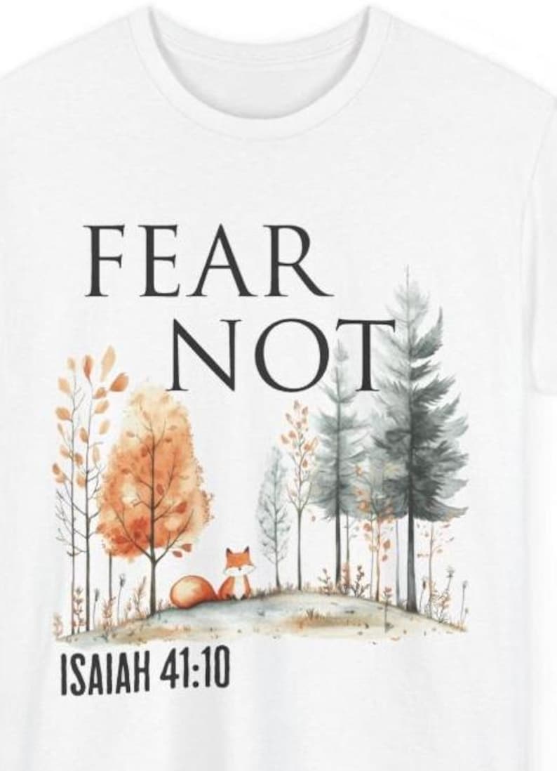 May include: A white t-shirt featuring a watercolor-style graphic of a small orange fox sitting in a forest with orange and grey trees. The text "FEAR NOT" is printed in black above the forest, and "ISAIAH 41:10" is printed in black below the fox.