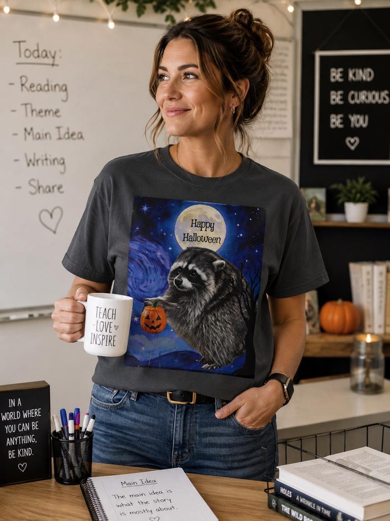 Op de afbeelding: Een antracietgrijs T-shirt met ronde hals en een afbeelding van een wasbeer die een pompoen vasthoudt onder een volle maan met de tekst Happy Halloween. Het shirt wordt gecombineerd met een witte mok met de tekst TEACH LOVE INSPIRE.