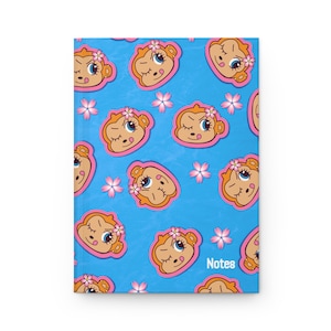 May include: A blue notebook cover featuring a repeating pattern of cartoon monkey faces with pink flower accents. Each monkey has a winking expression with a tongue sticking out. The word Notes is printed in white in the bottom right corner.