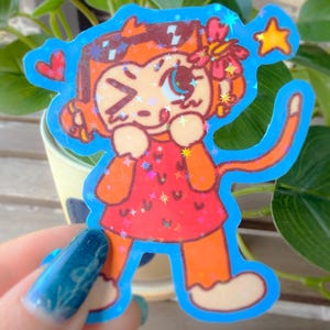 May include: A holographic sticker featuring a cartoon monkey with orange fur, wearing a red dress with a dark pattern. The monkey has a flower in its hair, a long tail, and is winking. The design includes a red heart and a yellow star on a blue background.