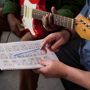 Può includere: Una chitarra elettrica rossa e bianca è tenuta da una persona, mentre un'altra persona indica un diagramma di accordi con una penna blu. Il diagramma è un quaderno a spirale con vari diagrammi di accordi per chitarra.