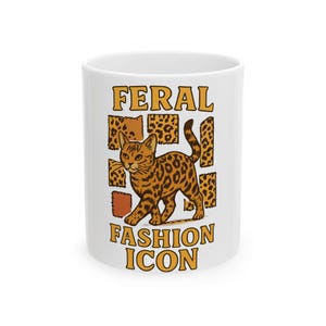Peut inclure: Mug en céramique blanche avec une illustration de chat tacheté et les mots "FERAL FASHION ICON" en orange. Le chat est brun avec des taches noires, et le design comprend des carrés à imprimé léopard.