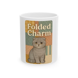 May include: White ceramic mug with a patchwork design, the words "Folded Charm", and a gray Scottish Fold cat illustration. The mug has a rounded shape and a simple handle.