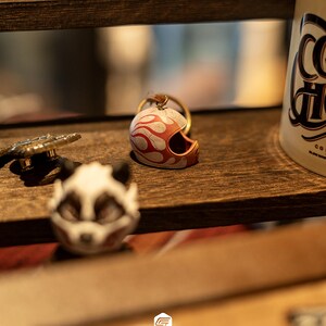 May include: A miniature motorcycle helmet keychain featuring a cream-colored base with orange flame graphics. The helmet has a gold-toned metal ring attached to the top, and it is positioned on a dark wooden shelf next to other small objects.
