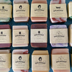 May include: A collection of 12 bars of handmade soap with brown paper labels. The labels feature the text "Speakeasy Soap Co." and various scents, including "Rosemary Mint", "Lemongrass", "The Classic Bar in Town", "The Best Tasting Tea Bar", "The Sweetest Victorian", "Black Tea & Bergamot", "Lavender Thyme", "The Classic Bar in Town", "The Classic Bar in Town", "The Sweetest Victorian", "The Best Tasting Tea Bar", and "The Classic Bar in Town".