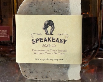 May include: A bar of Speakeasy Soap Co. soap with a light beige label. The soap is a light gray color with dark flecks. The label features a drawing of a person bending over and the text "Speakeasy Soap Co."