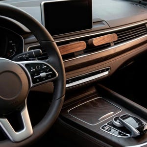 May include: Two rectangular dark wood car air freshener clips attached to the horizontal slats of a vehicle dashboard vent. The interior features a black leather steering wheel, a digital display screen, and dark wood trim accents on the console.