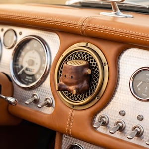 May include: A small, dark brown wooden figure with two black eyes and two small light-colored feet, shaped like a tree stump, mounted inside a circular brass-rimmed car air vent on a tan leather dashboard with silver engine-turned metal panels.