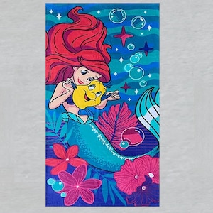 Puede incluir: Una toalla de playa rectangular con una colorida ilustraci&oacute;n de Ariel de La Sirenita. Ariel tiene el pelo rojo y una cola azul, sosteniendo un pez amarillo. El fondo incluye burbujas, estrellas y flores rosas y rojas.