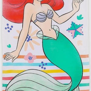 May include: A white beach towel featuring a cartoon illustration of a mermaid with red hair and a green tail. The towel has a striped pattern in various colors, with floral and starfish accents. The mermaid is wearing a seashell bra.
