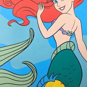 Puede incluir: Ilustraci&oacute;n de Ariel, la Sirenita, con cabello rojo ondeante, ojos azules y una cola verde. Un pez amarillo y azul, Flounder, nada cerca. El fondo es un degradado de azul.