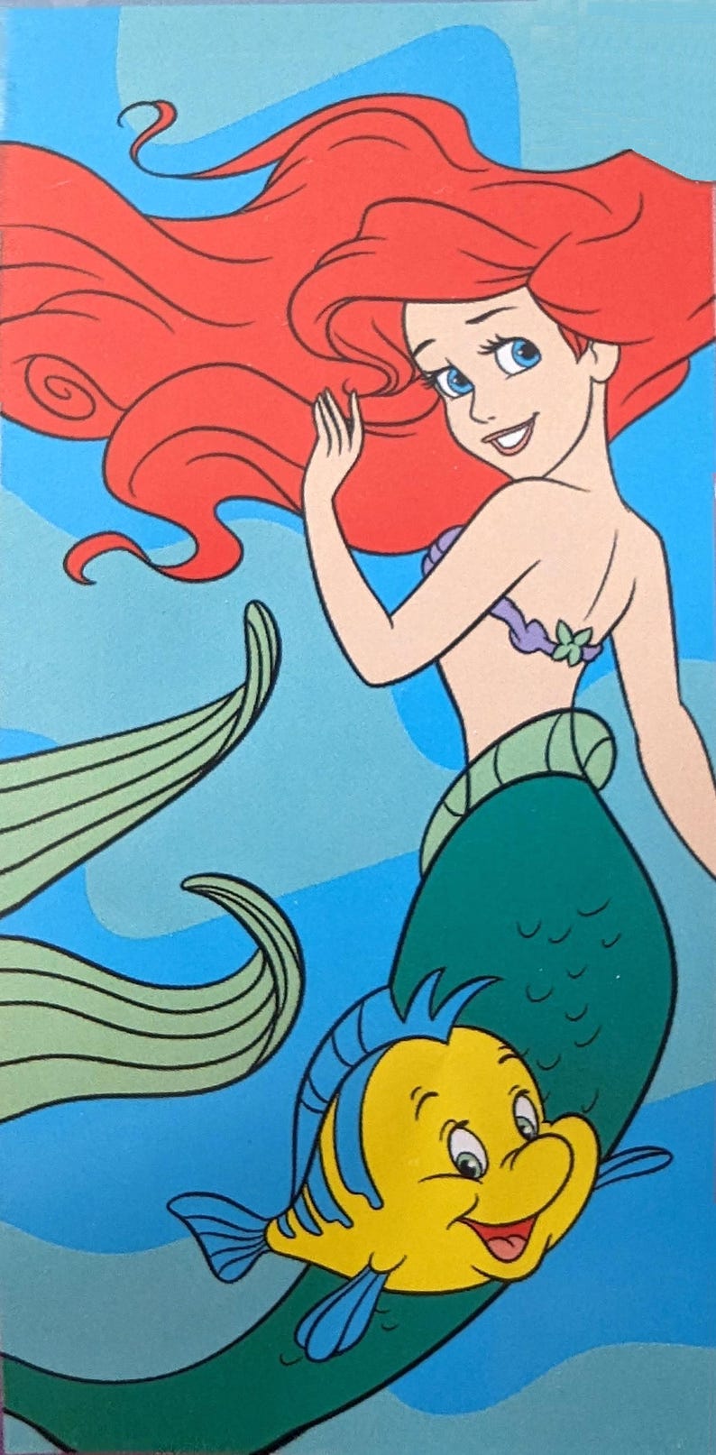 Puede incluir: Ilustraci&oacute;n de Ariel, la Sirenita, con cabello rojo ondeante, ojos azules y una cola verde. Un pez amarillo y azul, Flounder, nada cerca. El fondo es un degradado de azul.