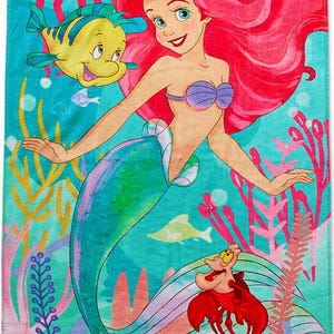 May include: A beach towel featuring a colorful illustration of Ariel, the Little Mermaid, with Flounder and Sebastian. The design includes a turquoise background with coral, fish, and other sea life. Ariel has red hair and a purple seashell bra.