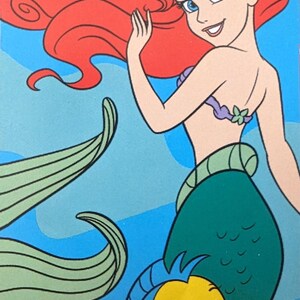 Puede incluir: Ilustraci&oacute;n de Ariel, la Sirenita, con cabello rojo ondeante, ojos azules y una cola verde. Un pez amarillo y azul, Flounder, nada cerca. El fondo es un degradado de azul.