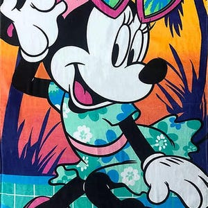 Puede incluir: Toalla de playa con Minnie Mouse en una escena veraniega. Minnie lleva un vestido floral, gafas de sol en forma de coraz&oacute;n y sandalias rosas. El fondo muestra palmeras y una puesta de sol.