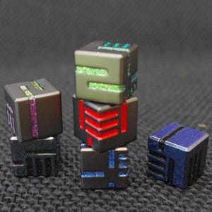 May include: A collection of five small, metallic, cube-shaped dice. One die is blue, while the others are dark gray with recessed grooves filled with glittery paint in shades of green, purple, red, and blue. The dice feature geometric patterns.