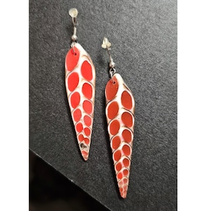 May include: A pair of dangle earrings with a red and white pattern. The earrings have a teardrop shape with a series of red ovals on a white background. The earrings are made of a lightweight material and have silver-colored hooks.