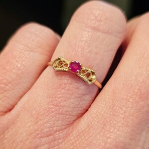 May include: A gold ring with a small, round, red gemstone at the center. The ring has an ornate design on either side of the gemstone. The ring is on a finger.
