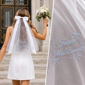 May include: A white tulle wedding veil featuring a large satin bow at the top and light blue embroidery that reads Future Mrs. Bennett, accented with small embroidered bows and hearts on the sheer fabric.