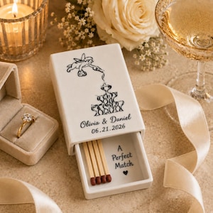 May include: A white ceramic sliding matchbox featuring a black line drawing of a cherub pouring champagne into a tower of glasses. The text reads Olivia & Daniel 06.21.2026 on the exterior and A Perfect Match with a heart inside the tray.